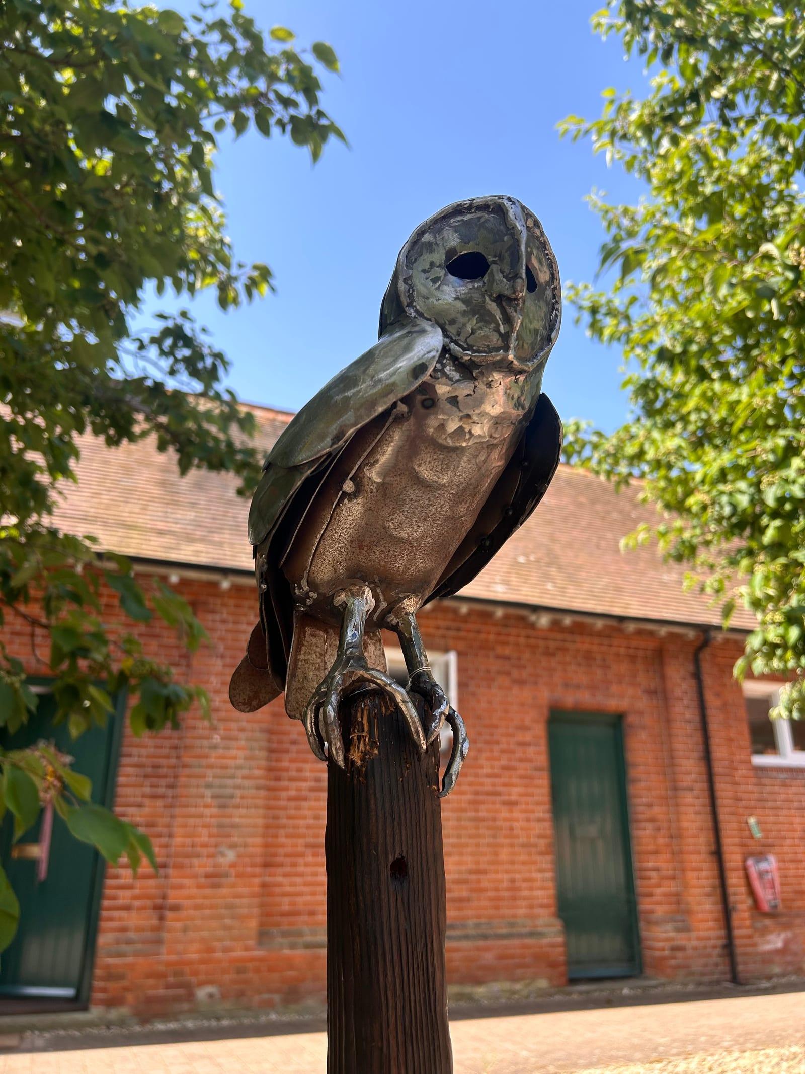 Steel barn owl sculpture in weathered mild steel, its surface a mottled grey-green and rust patina, perched on a wooden post against a red-brick building and trees.