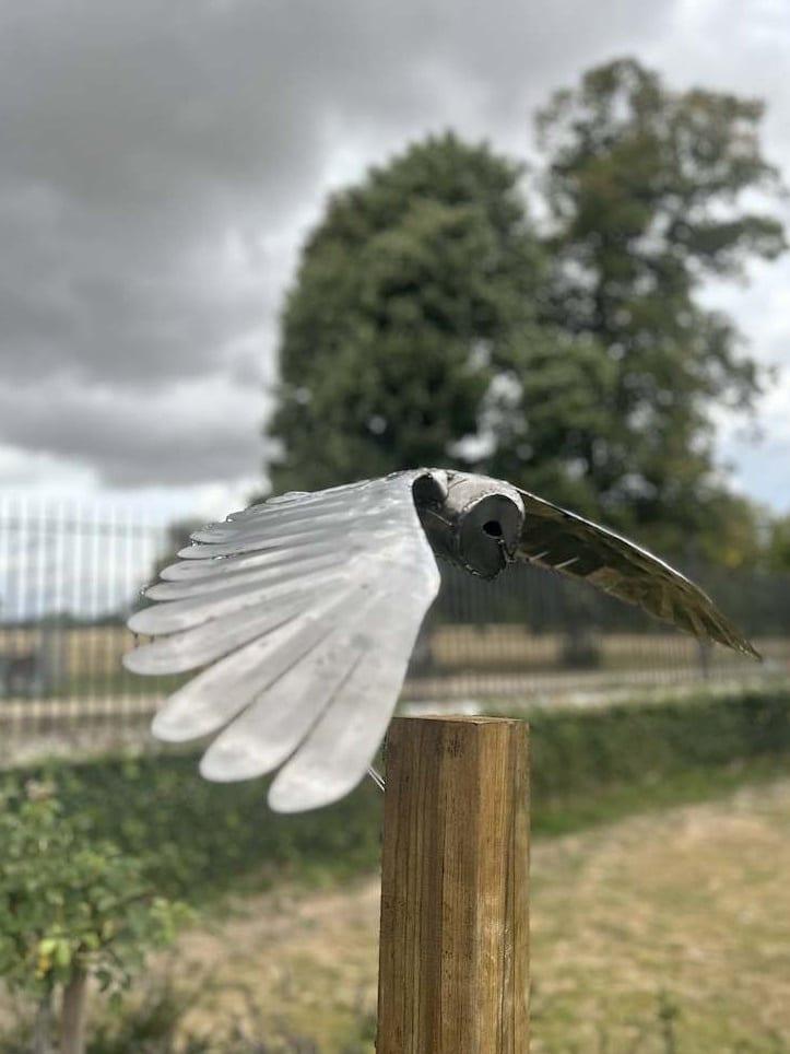 Steel barn owl sculpture in polished stainless steel, wings outstretched level in gliding flight, mounted on a wooden post, a railed parkland and grey sky behind.