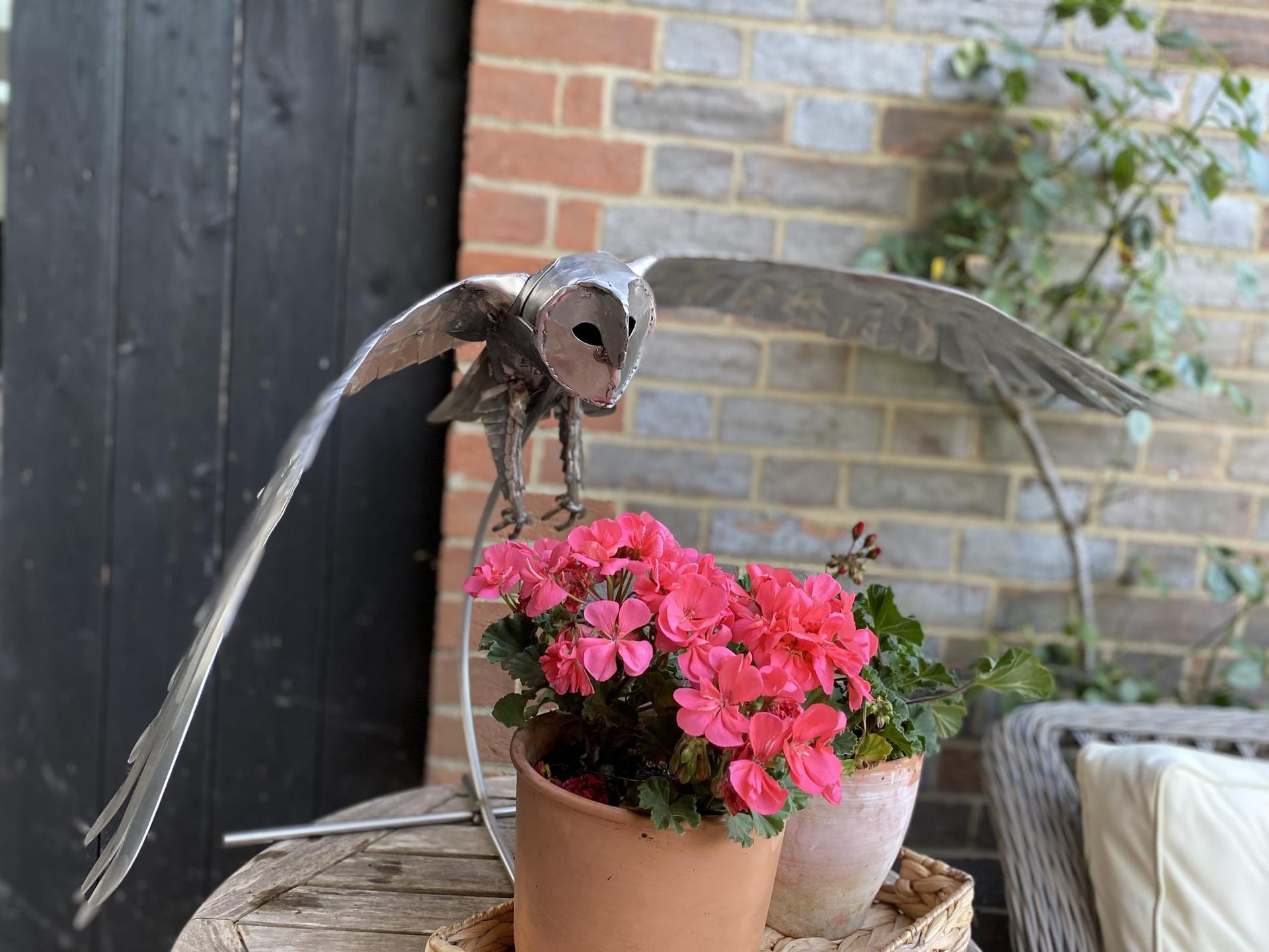 Steel barn owl sculpture in polished stainless steel, wings swept back in a shallow dive toward a pot of pink geraniums, a brick wall behind.