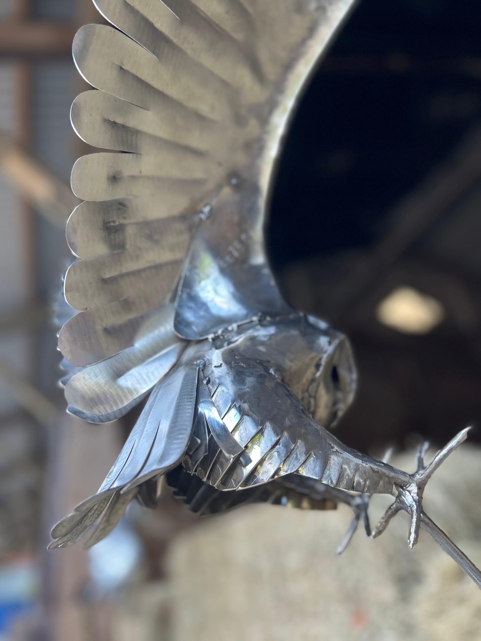 Close view of the stainless steel barn owl mid-strike, its raised wing and hand-formed face disc filling the frame, in a barn interior.