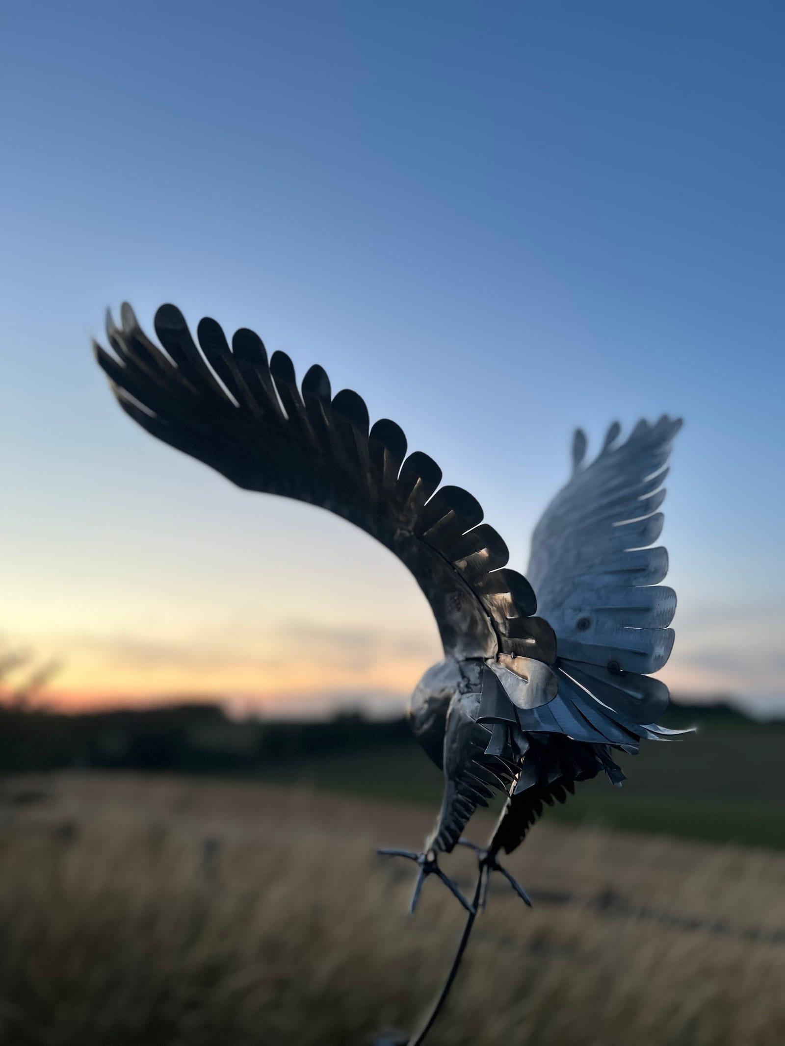 Steel barn owl sculpture striking with wings raised, in stainless steel, silhouetted on a curved rod against a dusk sky over open fields.