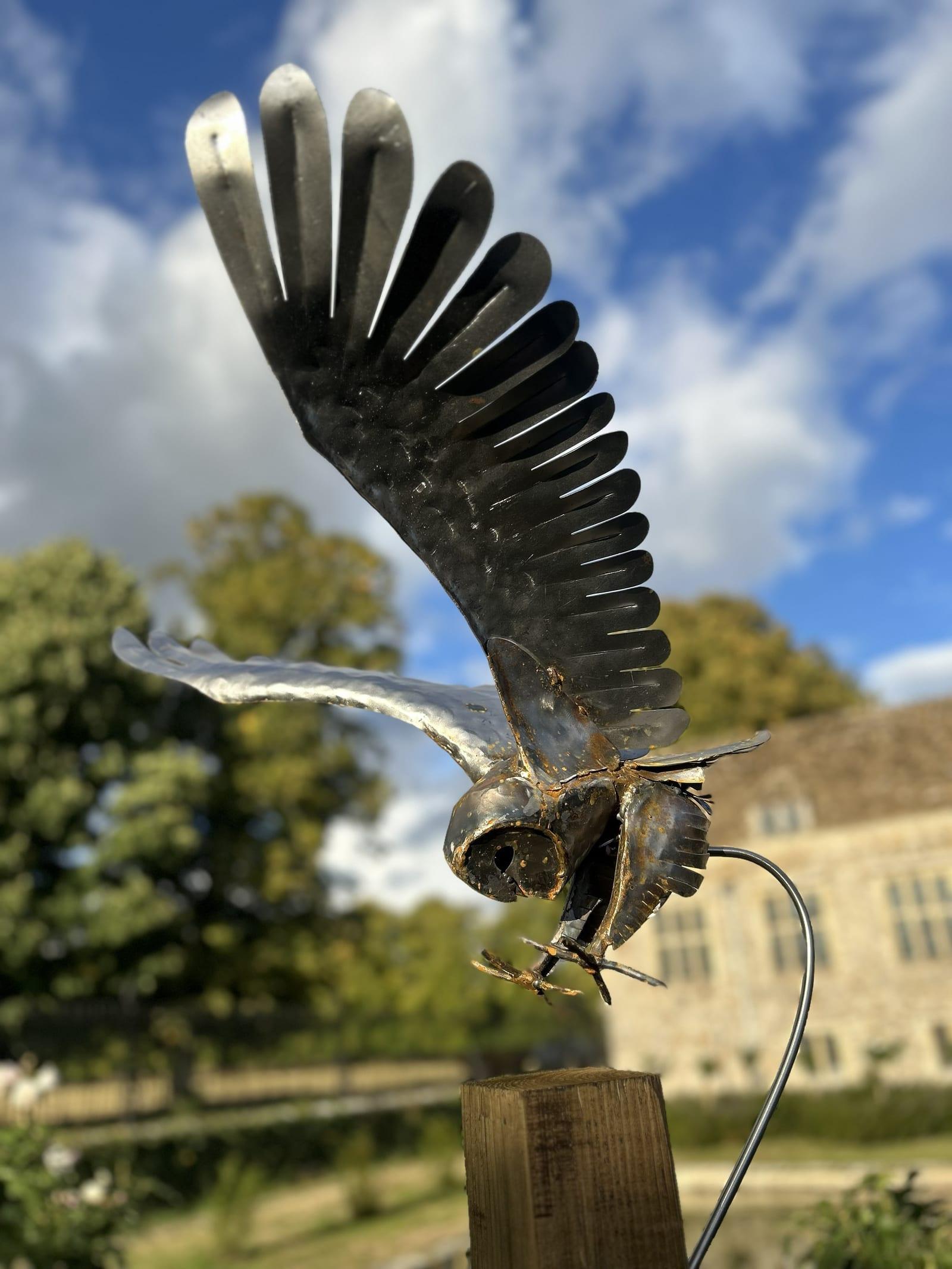 Steel barn owl sculpture in darkened mild steel with light rust, wings raised mid-strike, mounted on a wooden post with a curved steel arm, a stone manor house behind.